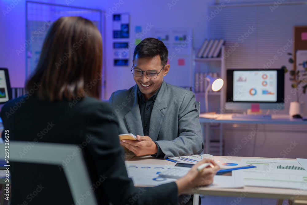 Poster two businesspeople are working late in the office, discussing over some paperwork and using a smartp