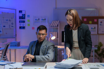 Two business professionals engage in document analysis at a modern office desk. Charts and graphs adorn the walls, suggesting a focus on data-driven decision-making and teamwork