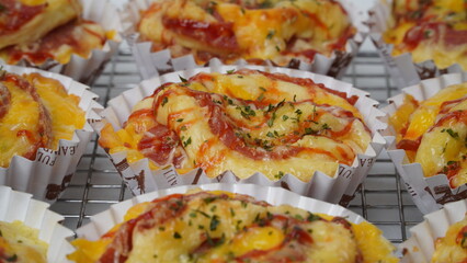 A group of salty bread is served in a paper cup with smoked beef filling, orange cheddar and topped with mayonnaise, tomato sauce, and parsley which is placed on a cooling rack with a white background