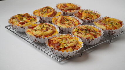 A group of salty bread is served in a paper cup with smoked beef filling, orange cheddar and topped with mayonnaise, tomato sauce, and parsley which is placed on a cooling rack with a white background