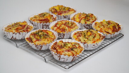 A group of salty bread is served in a paper cup with smoked beef filling, orange cheddar and topped with mayonnaise, tomato sauce, and parsley which is placed on a cooling rack with a white background