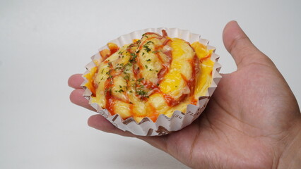 A single salty bread is served in a paper cup with smoked beef filling, orange cheddar and topped with mayonnaise, tomato sauce, and parsley which is on hand with a white background