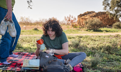 Fototapeta premium Young woman sitting on a picnic blanket in nature, holding two metal mugs while searching through her bag, with hiking shoes and a notebook nearby in a peaceful rural setting