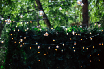 Blurred image of light bulbs outdoor on a wire against dusk forest, holiday concept. Outdoor decor, dark festive background with a copy space