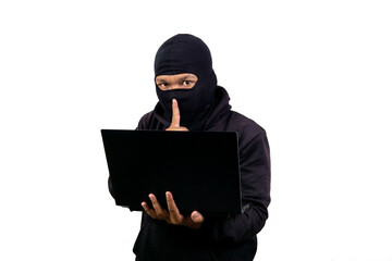 Male hacker in mask and black jacket puts finger in front of lips gesturing stay still or keep...