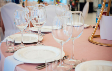Table setting with light pink tablecloth close up. Wine and beverage glasses, white plates, forks and knives, preparation of the restaurant for the reception of guests