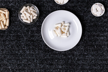 Dietary supplements, many vitamins and minerals in small plates and jars on black background from above. Big amount of white colored pills and nutritional supplements, top view. Healthy lifestyle