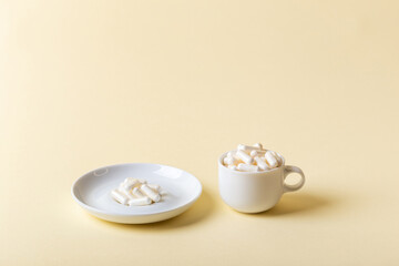 White ceramic cup and white plate of white pills. Vitamins and minerals, dietary supplements on beige background. Minimalist composition with white capsules.