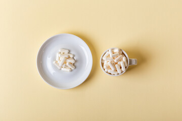 White ceramic cup and white plate of white pills top view. Vitamins and minerals, dietary supplements on beige background from above. Minimalist composition with white capsules.
