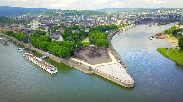 Aerial view of Deutsches Eck in Koblenz, Germany, where the Rhine and Moselle rivers merge. The historic monument, lush greenery, and passing cargo boats create a dynamic and scenic riverside scene.