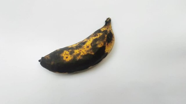 A yellow banana that is starting to blacken its skin on cutting board with a white background. The blackening of the banana skin is a sign that the banana is overripe but is still edible.