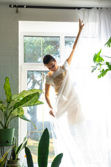 Woman in apron hangs transparent tulle curtains on large windows in the house inside the interior with potted plants. Spring cleaning, tidying up