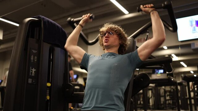 A man is performing a shoulder press exercise using gym equipment. The workout focuses on building upper body strength, with the gym's modern atmosphere enhancing the fitness experience.