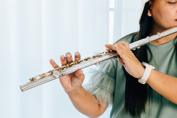 A green shirted woman plays a silver flute while sitting on a brown couch at home. The living room warm tones enhance the peaceful and expressive mood of her musical performance.