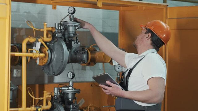 A male engineer in a safety helmet and uniform with a digital tablet in his hands inspects a gas distribution system. An engineer checks the safety of pipe connections.