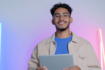 Young latin student smiling and holding tablet with neon lights background