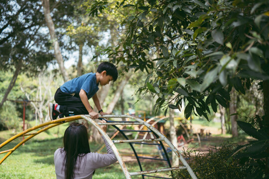 A young 8 years old boy climbing a jungle gym with the assistance of his mother in a park, surrounded by green trees and natural light.