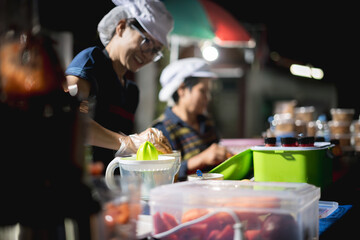 Thai woman vendor smiling while preparing juice at a night market, with another vendor working in the background.