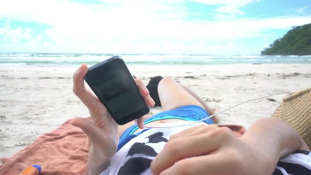 A woman is laying on the beach with her feet up and a cell phone in her hand. The beach is empty and the sky is cloudy