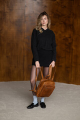 A girl in a warm sweater, a black short skirt, nylon tights, and black shoes is posing with a brown backpack.
