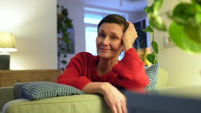Mature woman smiles and laughs happily at camera, sits comfortably on sofa. Female watch at family member, warmth and affection, shared happiness and emotional support, look of unconditional love