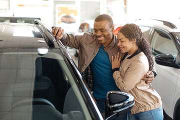 Car Choosing. Happy African Spouses Standing Next To Modern Vehicle In Showroom, Cheerful African American Couple Purchasing New Automobile In Dealership Center, Discussing Auto Characteristics. High