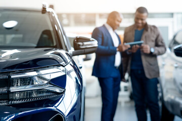 Black Male Customer In Car Dealership Center Looking At Tablet With Salesman, Happy African...