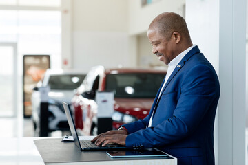 Positive black car salesman using laptop computer at work desk in auto dealership. Cheerful African...