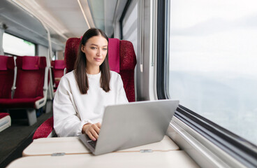 Portrait of young female freelancer working online on laptop while traveling on train, sitting near...
