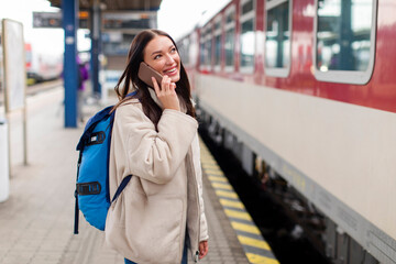 Cheerful lady traveler talking on cellphone at railway station platform, waiting for boarding to train wagon, ready for weekend trip