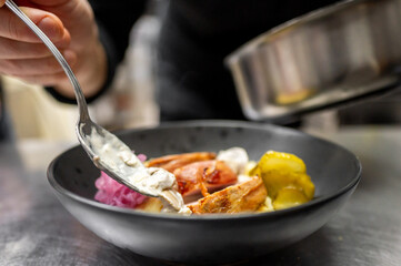 A close-up of a hand serving a dish in a black bowl. The meal features tender chicken pieces, sauce, pickles, and fresh onions, highlighting a culinary presentation in a modern kitchen setting.