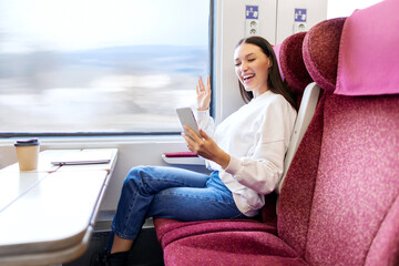 Happy lady blogger making video call on smartphone while sitting in train near window, woman waving to camera while talking online in train wagon