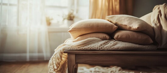 Soft beige plush pillows stacked on a wooden bench in a sunlit room with sheer curtains and softly blurred decorative elements in the background