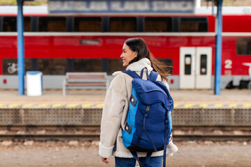 Obraz premium Tourist woman looking aside at arriving train, standing at railroad station platform, wearing backpack, travelling through Europe by train, back view