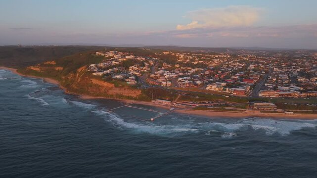 Drone tracking left of Merewether Ocean Baths, panning to reveal Newcastle&rsquo;s urban skyline at golden hour sunset