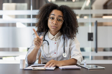 Portrait of a serious young African American female doctor sitting in a hospital office at a table with documents, talking and gesturing to the camera