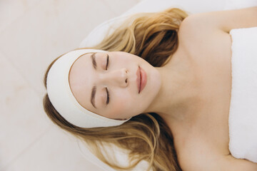 Fototapeta premium Blonde woman with headband lying with closed eyes on massage table during skincare treatment at spa