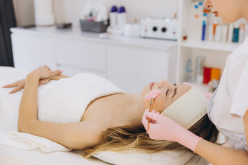 Cosmetologist applying a pink mask on the face of a young woman lying on a massage table in a beauty salon
