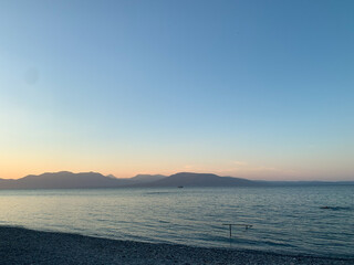 Calm sea with distant mountains under a clear sky at sunset. Gentle waves reach the pebble shore....