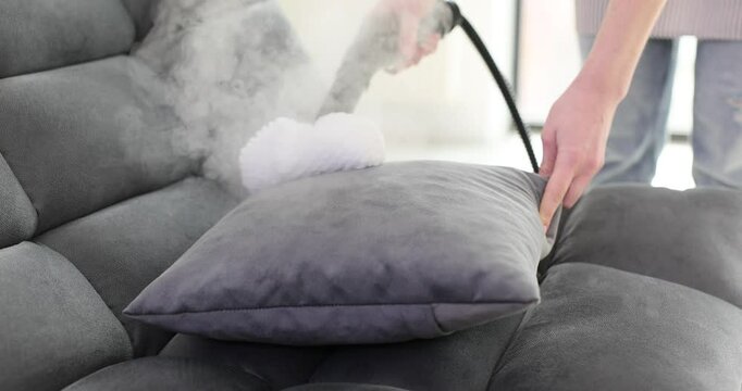 Cleaning service worker steams pillow on sofa in office. Woman eliminates dust mites and bacteria using specialized equipment for home hygiene