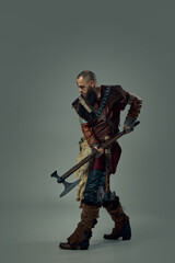 Viking warrior gripping double-headed axe, crouching in battle-ready stance, wearing fur cloak and rugged armor against dark studio background. Concept of medieval battlefield, warrior spirit.