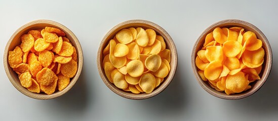 Assorted yellow vegetable crisps in three round bowls arranged symmetrically on a light gray surface showcasing healthy snacking options.