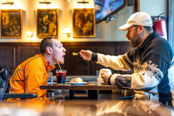 Man feeding disabled friend in restaurant, showing support and care