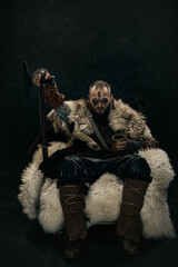 Viking warrior seated on fur-covered throne gripping axe and drinking from wooden mug, dressed in leather and fur against dark background. Concept of historical portrait, Scandinavian culture.