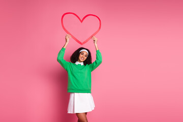 Cheerful woman holding a red heart shape on pink background wearing green jumper and white skirt