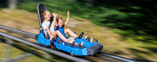 Pair of cute laughing kids enjoy have fun riding alpine roller coaster cart together at Harz nature...