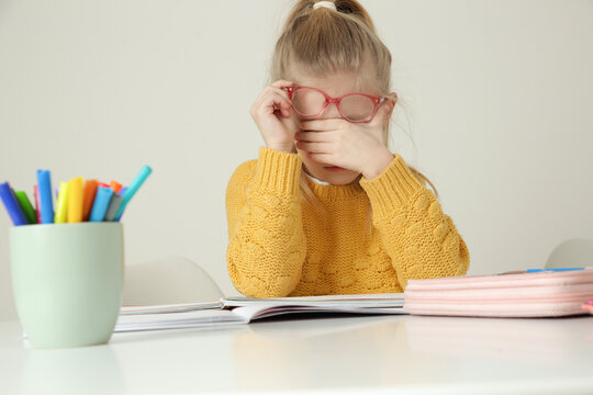 Tired elementary school girl with eyeglasses rubbing her eyes while doing homework