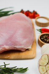 Vertical close-up photo of side view of a piece of turkey fillet brisket lying on a wooden cutting board on a white background surrounded by seasonings, herbs and vegetables. The concept of healthy