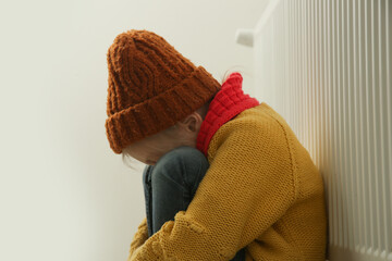 Little girl in woolen winter clothes, near the radiator of the home heater trying to keep warm. Restrictions and savings of gas and electricity during energy or economic crisis, rising prices.	