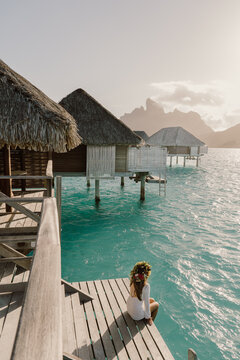 A woman in a white dress and floral crown sits by overwater bungalows, gazing at the turquoise lagoon in Bora Bora. A dreamy escape to paradise, where tranquility meets breathtaking natural beauty